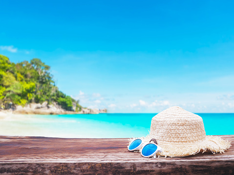 Close up straw hat and sunglasses on wooden table over blurry background of summer beach with blue sky and white sand landscape.