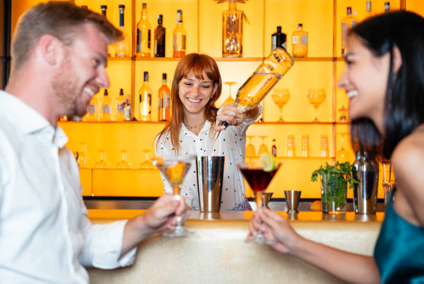 barkeeper gießt alkohol aus der flasche in die gläser - glückliches schönes paar, das am wochenendabend an der cocktailbar in der hotellobby abhängt - lifestyle-konzept mit barmann, der getränke zubereitet und kunden bedient - nightlife party group of people martini stock-fotos und bilder