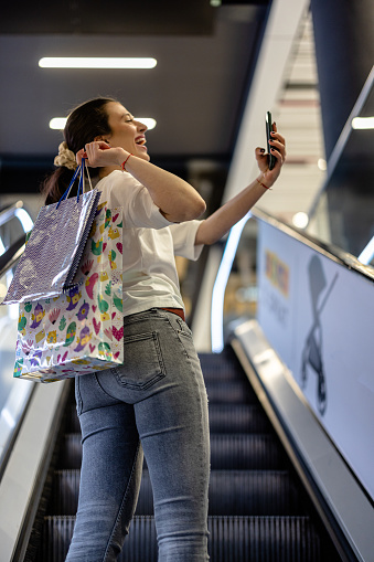 A young cheerful woman taking selfie while moving up with escalator in shopping mall