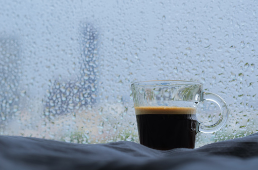 A glass of Espresso coffee shot puts on bed with rain drop on window in raining day. Stay home concept.