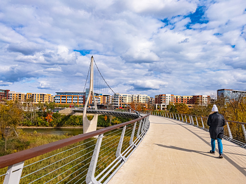 A woman is walking across a pedestrian and bicycle bridge in the fall. The modern office and apartment buildings of downtown Dublin Ohio are in the background. This elevated walkway is  the longest single-tower S shaped bridge in the world. The suspension tower with its cables is visible.  The bridge is over the Scioto River.