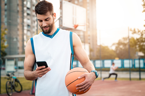 Portrait of smiling handsome male basketball player holding mobile phone and sports ball outdoors at sunset while listening to music over headphones.