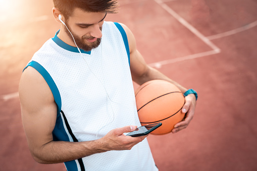 Athletic male basketball player listening to music while holding mobile phone and sports ball in. Sports guy enjoying healthy lifestyle outdoors and relaxing with music over smartphone.