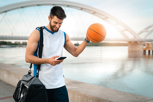 Handsome basketball player feeling happy and satisfied while looking at mobile phone walking by the river with sports equipment. Professional basketball player athlete using smart phone.