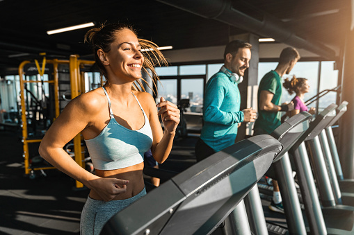 Happy athletic people jogging on treadmills in a health club. Beautiful people bathed in late afternoon sun running in a fancy gym. Selective focus of smiling slim fit woman exercising with friends in gym.