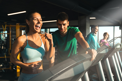 Young athlete male assisting a young woman while exercising on treadmills in a gym. Excited smiling girl running under her instructor's supervision.