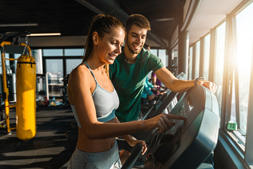 Happy athletic couple communicating while jogging on treadmills in a health club. Two good looking people having a fun during the gym workout. Supportive male fitness instructor with female gym client.