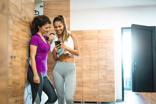 Two happy athletic women using mobile phone while standing in locker room and taking a break from exercising. Fit women after sports training with phone in locker room.