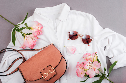 Feminine summer fashion flat lay with white blouse, bag, sunglasses and pink alstroemeria flower on grey background. Top view.