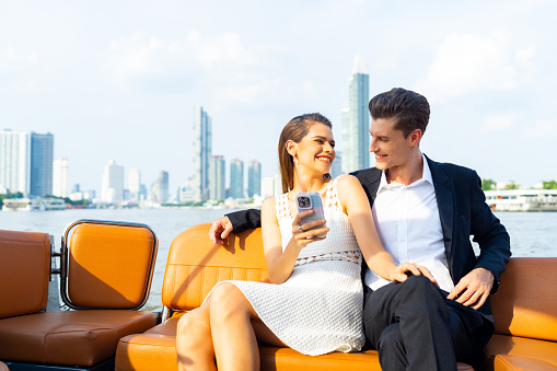 Happy Caucasian couple enjoy urban outdoor lifestyle using mobile phone together during travel city on luxury private boat yacht sailing in the river and celebrating holiday event at summer sunset.