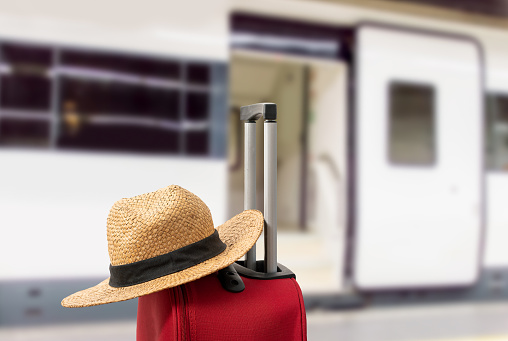 For travel and vacations purposes, a simple modern suitcase in red color at train station with a straw hat.