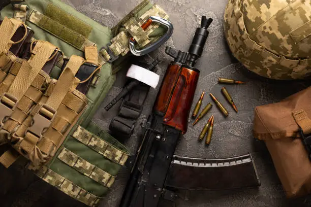 Photo of Military body armor, helmet and ammunition, a Kalashnikov assault rifle with matrons and a first aid tourniquet. On a black marble table. Army bulletproof vest. Flat lay. Military concept.