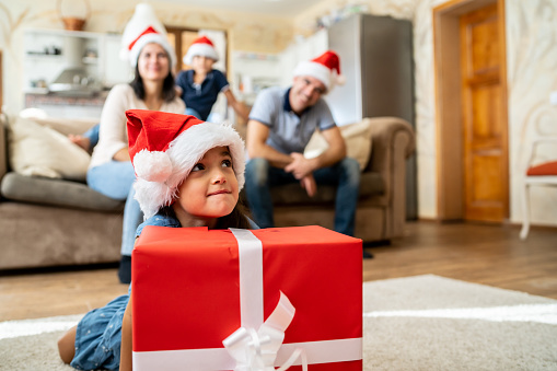 A young girl is wearing a santa hat and holding a large Christmas present, her parents and brother are sitting in the background also wearing santa hats
