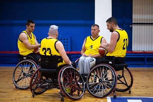 A circle of professional wheelchair basketball players sitting in their sports wheelchairs in basketball court, one man is holding the basketball