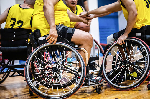 A group of unrecognizable disabled men sitting in customized professional basketball wheelchairs in a circle with outstretched arms and stacking their hands at indoor sports hall