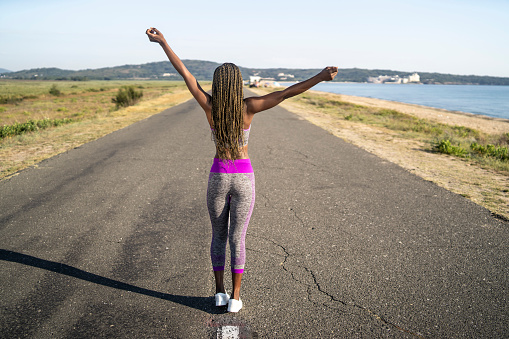 A rear view of an African-American woman walking in the middle of an empty highway next to a coastline wearing modern running leggings and t-shirt under a clear blue sky with her arms outstretched in the air