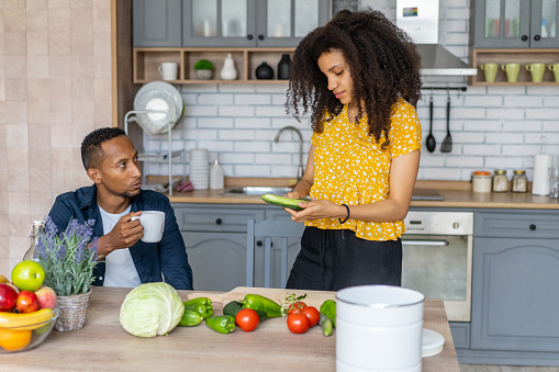 A woman is standing and peeling vegetables for a meal in a bright modern kitchen, her boyfriend is watching her while he drinks from a white mug