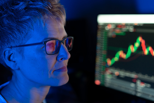 A serious looking woman with short hair and eye glasses looking away from camera, with a graph illuminted on a screen beside her against a dark background