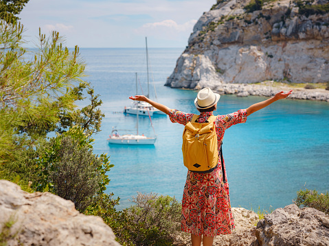 Asian woman in hat look on views of azure Bay in Mediterranean sea. Travel and vacation concept. Anthony Quinn bay with crystal clear water in Rhodes island, Greece. The most beautiful beach.