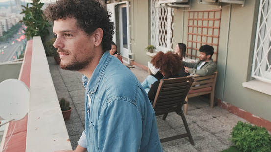 Close up, young guy looks at the city standing on the balcony, his friends are chatting in the background