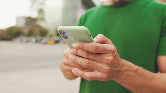 Close-up of mature man's hands wearing casual clothes standing on modern buildings background, scrolling on smartphone