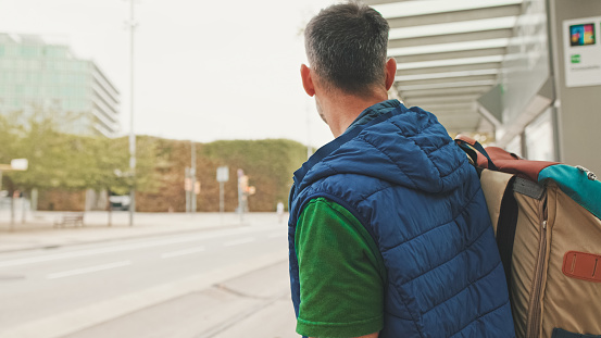 Middle-aged man dressed in casual clothes, with backpack on his shoulder, looks around while waiting for tram at public transport stop in the city