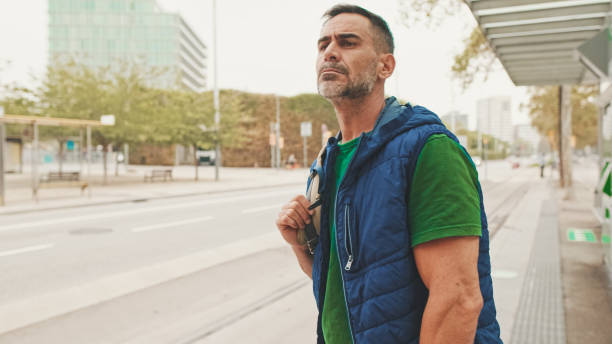 istock Middle-aged man dressed in casual clothes, with backpack on his shoulder, looks around while waiting for tram at public transport stop in the city 1528145135