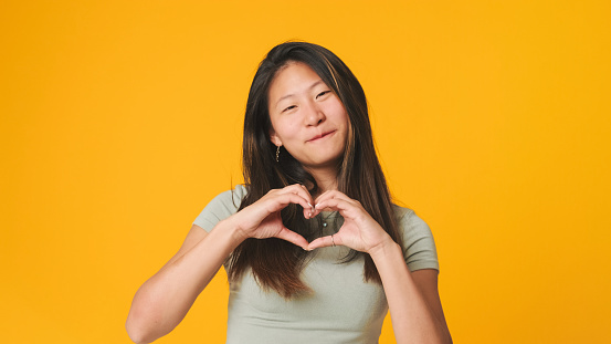 Girl in gray top looking at camera, with smile making heart shape with hands, expressing love care isolated on yellow background in studio