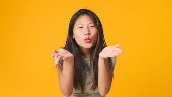 Girl in gray top looking at camera with smile, blowing lots of air kisses isolated on yellow background in studio