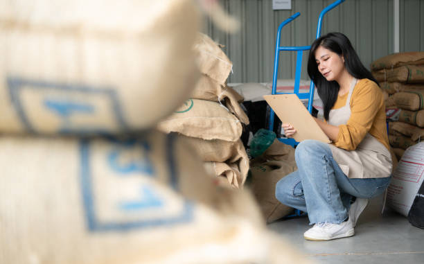 junge asiatische frau, die im kaffeebohnenlager auf dem boden sitzt und dokumente überprüft - coffee bag green bean stock-fotos und bilder