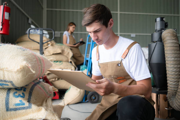 portrait of a young male worker in overalls working checking coffee beans bag in a coffee beans warehouse - coffee bag green bean imagens e fotografias de stock