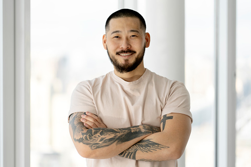 Cheerful, smiling Asian man crossed his arms. An attractive man has stylish tattoos on his hands, standing in a white office.