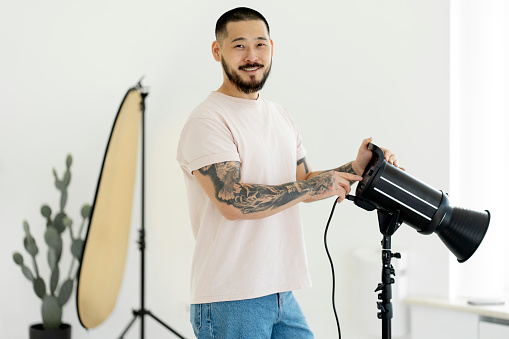 Portrait of professional Korean photographer with tattooed hands, holding camera equipment, adjusting light in photo studio. A smiling assistant in casual clothes prepares equipment for a photo shoot.