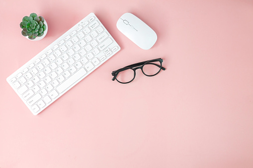 Female office desk with omputer keyboard, mouse, up of coffee, eyeglasses and green plant on pastel pink background