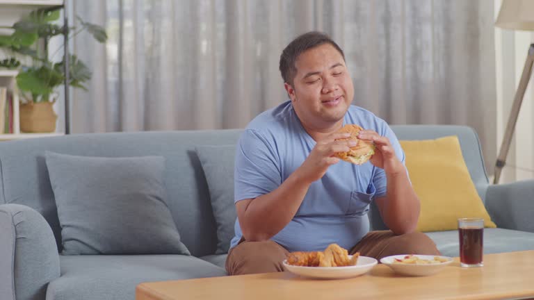 A Overweight Asian Man Smelling To Hamburger In His Hands Then Smiling While Eating Fast Food On A Sofa In The Living Room At Home