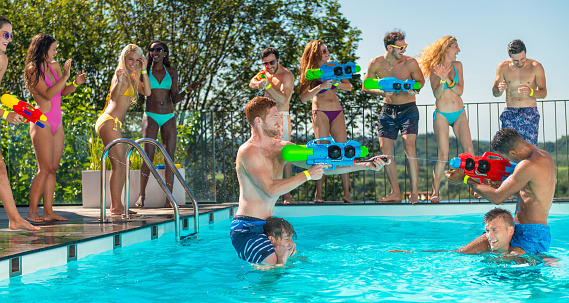 Two young men in the pool riding piggyback and spraying with squirt guns, young people in swimwear in the background. Holidays and lifestyle concept.