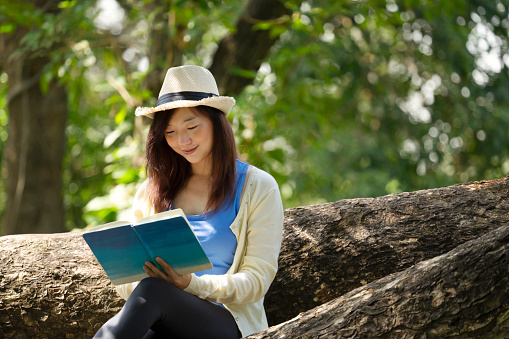young asian female wearing hat sitting on root of old big tree, peaceful attractive woman spend time  reading a book on summertime in the park