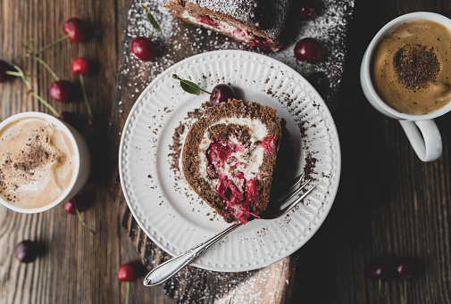 Cup of delicious korean dalgona coffee served with a fresh baked swiss roll with chocolate, cream and sour cherries on wooden background from above