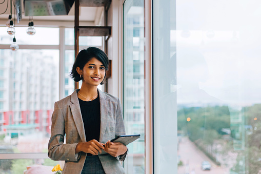 Waist up portrait of young female entrepreneur holding digital tablet and smiling happily at camera standing by window