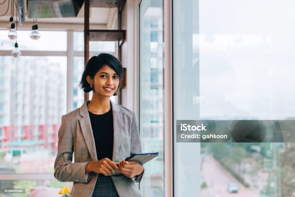 Young Businesswoman Posing by Window Waist up portrait of young female entrepreneur holding digital tablet and smiling happily at camera standing by window Businesswoman Stock Photo