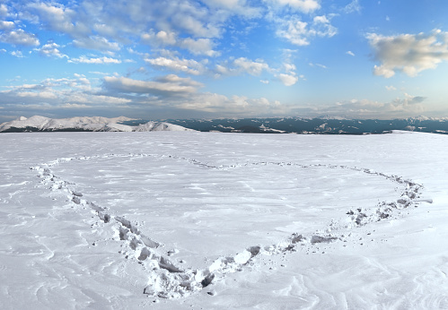 Human footprint form the heart shape on snow-covered mountainside plateau and mountain ranges behind.