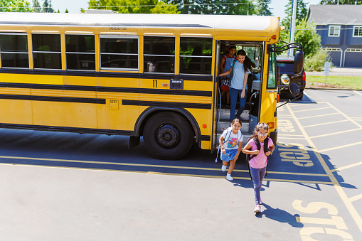 High angle view looking down at a multiethnic group of elementary age students arriving at school and disembarking their school bus which is parked in the school parking lot marked with a crosswalk.