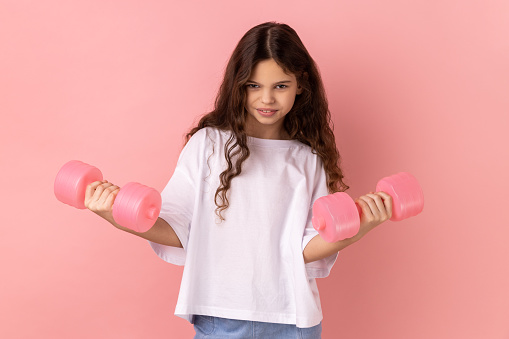 Portrait of optimistic little girl wearing white T-shirt holding pink dumbbells, training her arms, looking at camera with happy expression. Indoor studio shot isolated on pink background.