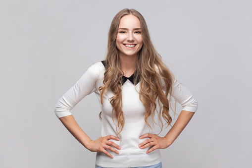 Portrait of happy joyful cheerful beautiful woman with long blond hair standing with hands on hips, looking at camera, expressing happiness. Indoor studio shot isolated on gray background.