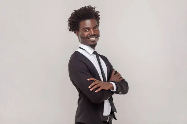 Portrait of cheerful man with Afro hairstyle with crossed arms, smiling broadly, laughing at good joke, wearing white shirt and tuxedo. Indoor studio shot isolated on gray background.