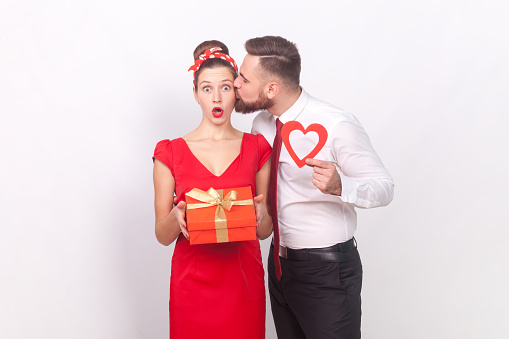 Shocked woman and romantic man in elegant clothes celebrating anniversary, husband showing red heart, wife looking at camera with amazed face. Indoor studio shot isolated on gray background.