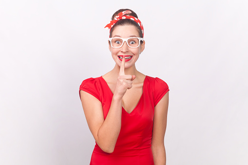 Portrait of funny happy smiling woman wearing red dress and head band keeps hand near lips, asking to keep secret, private information. Indoor studio shot isolated on gray background.