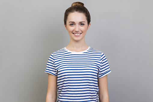 Portrait of attractive charming woman wearing striped T-shirt standing looking at camera with toothy smile, expressing happiness and positive emotions. Indoor studio shot isolated on gray background.