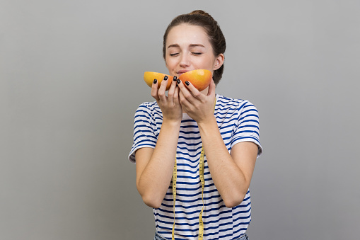 Portrait of woman wearing striped T-shirt with measure tape on shoulders, enjoying smell of delicious grapefruit, concept of healthy eating. Indoor studio shot isolated on gray background.