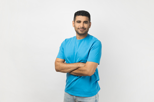 Portrait of handsome joyful unshaven man wearing blue T- shirt standing looking at camera, expressing positive emotions, keeps arms crossed. Indoor studio shot isolated on gray background.